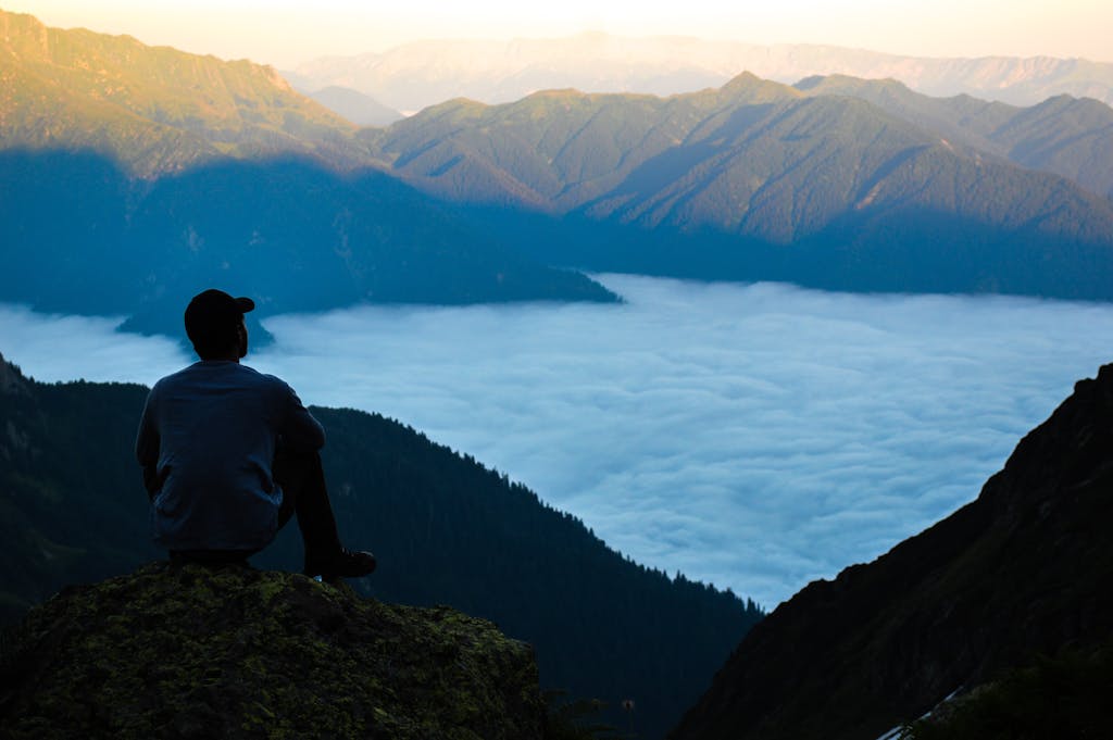 Man sits on mountain peak overlooking ranges and clouds, shadowy mountains in foreground, distant peaks illuminated by sunlight