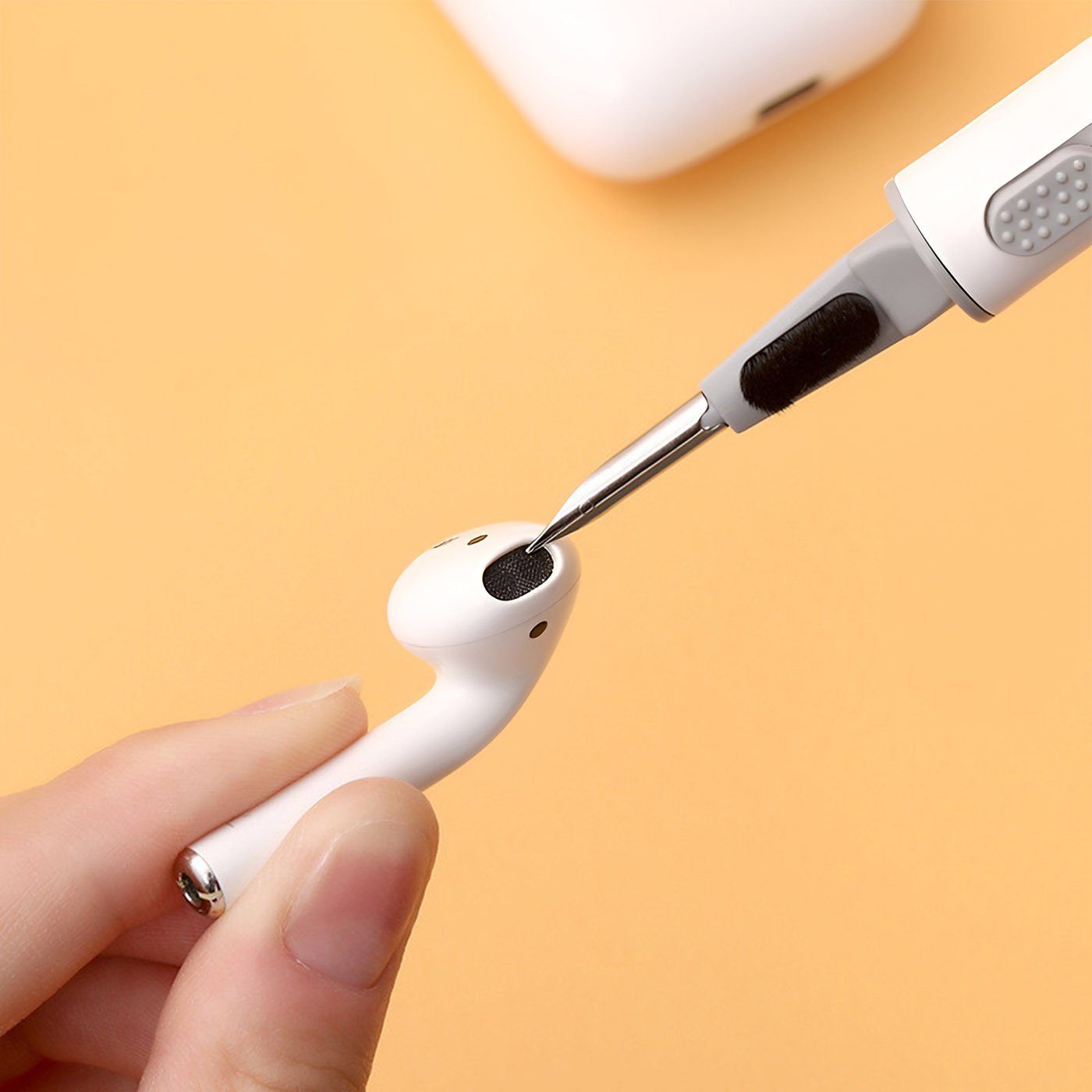 Metal tool cleaning mesh of white wireless earbud held between fingers on peach background.