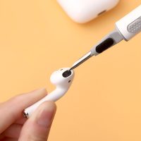 Metal tool cleaning mesh of white wireless earbud held between fingers on peach background.