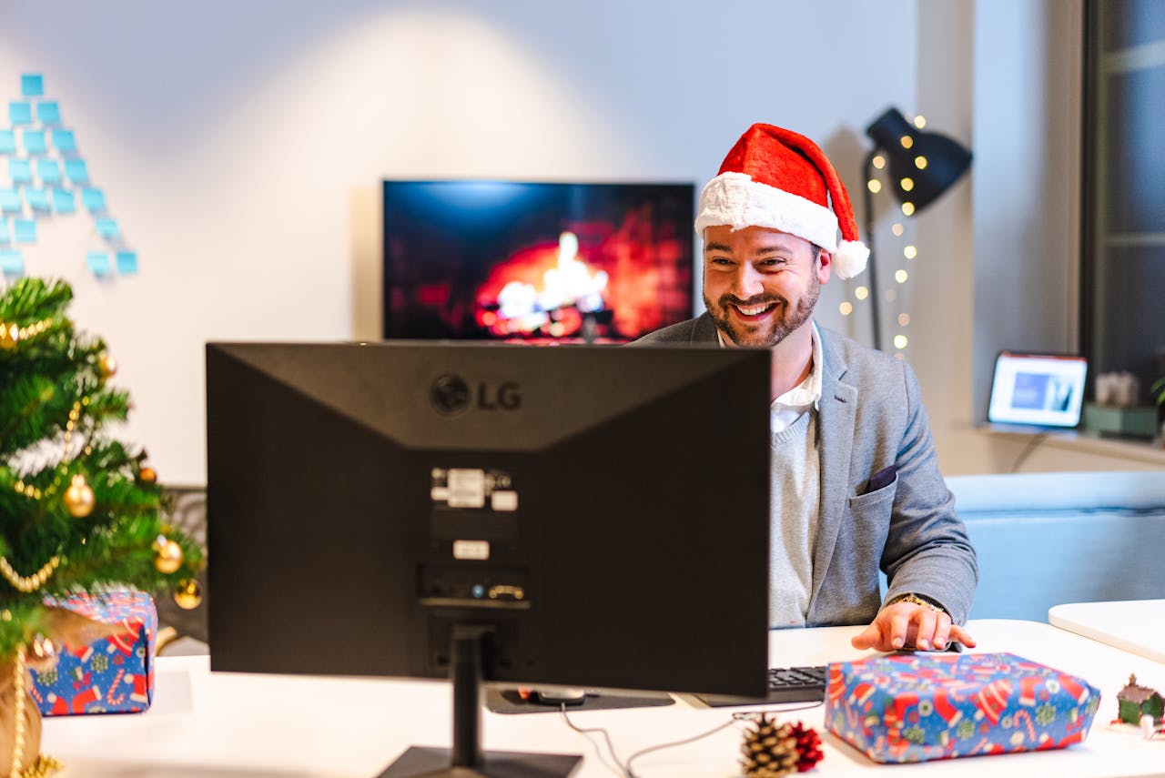 Man in Santa hat working at a festive setup with cozy desk accessories, a Christmas tree, and wrapped gifts.