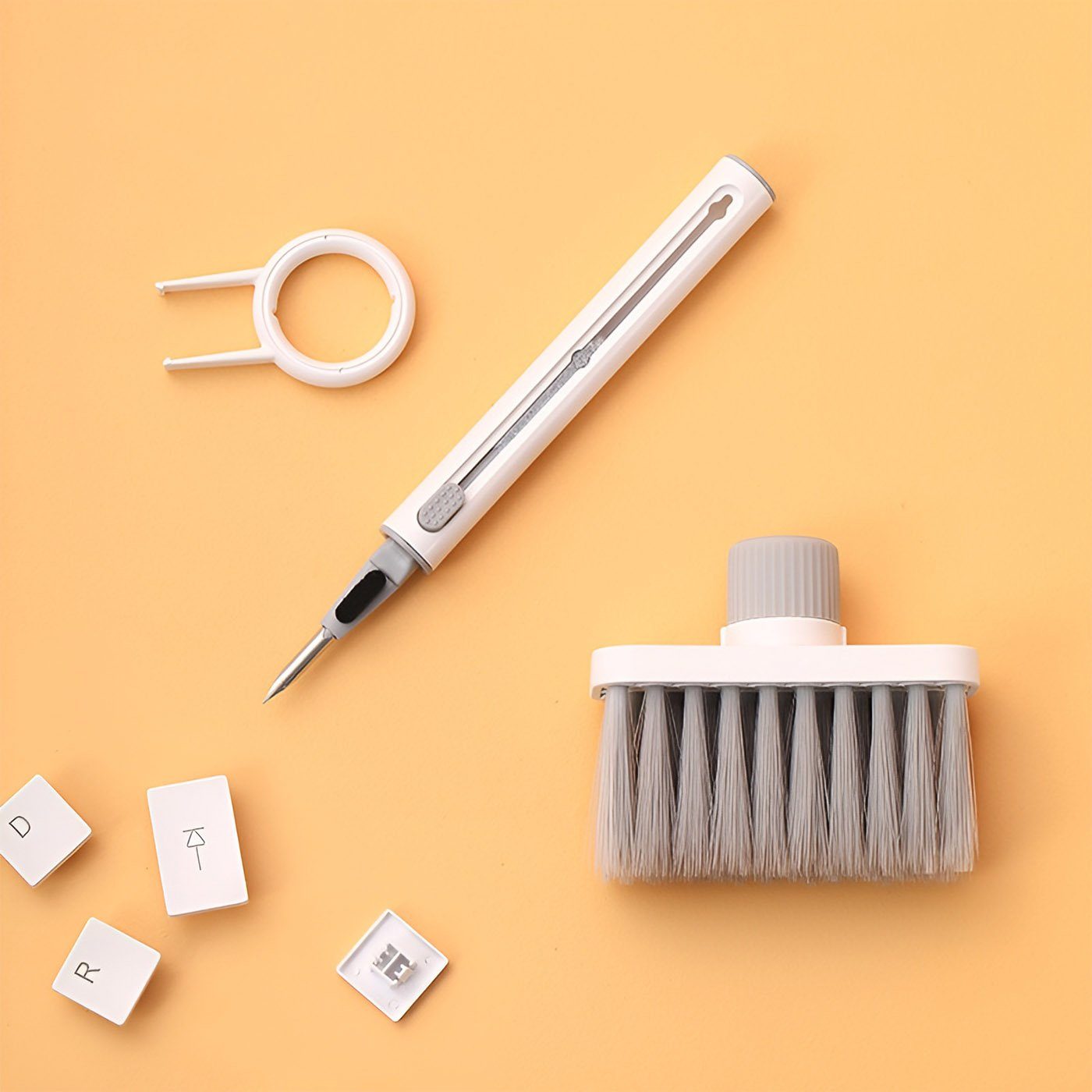 White keyboard cleaning kit with keyboard brush, earbud cleaning pen, and keycap puller, laid out next to keycaps.