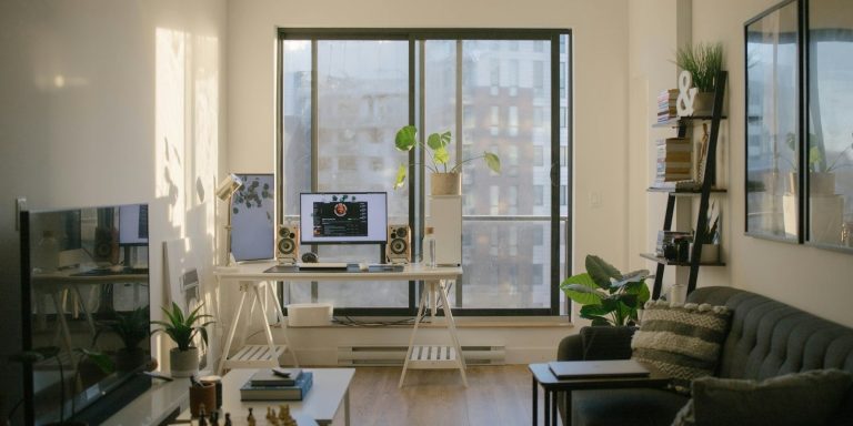 Bright living room with ergonomic home office setup, featuring desk, monitor, speakers, plants, and natural light.