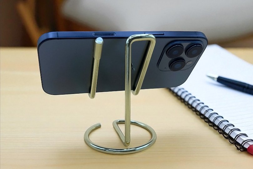 Serpentine metal phone stand Minimalist metal phone stand holding a smartphone horizontally on a wooden desk beside a notebook