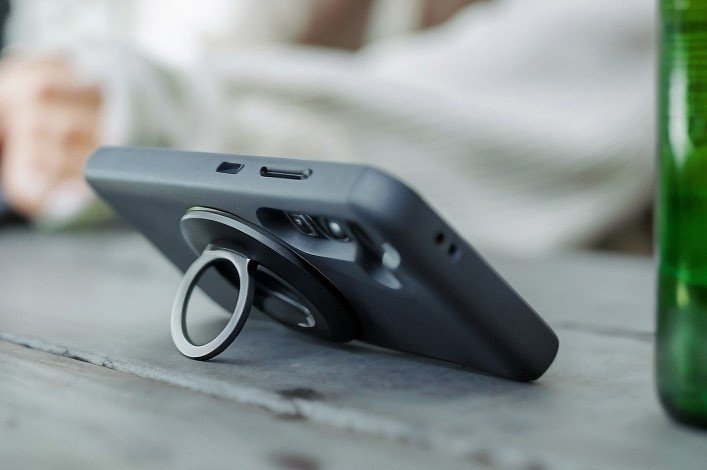 Beer-opener ring phone grip Smartphone with a black case and a metal ring grip attached, propped on a table outside next to a drink.