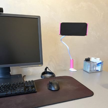 Pink flexible gooseneck phone holder clamped to an office desk, holding a smartphone next to a monitor.