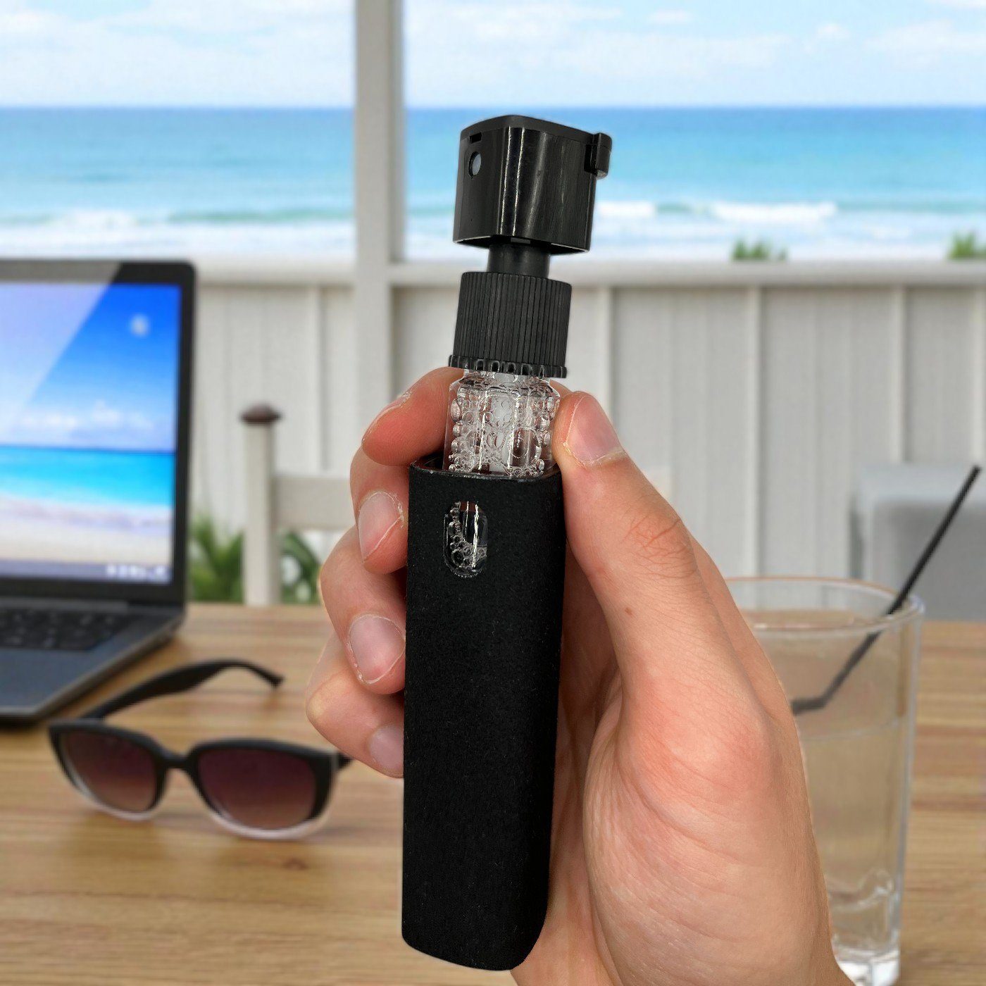 Hand holding screen cleaner with removable microfiber case at beachside workspace, with laptop and sunglasses.