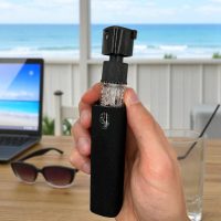 Hand holding screen cleaner with removable microfiber case at beachside workspace, with laptop and sunglasses.