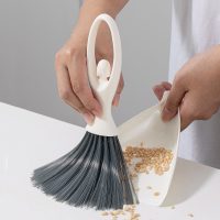 Hand sweeping food crumbs and grains off a table into the mini dustpan using the small brush.