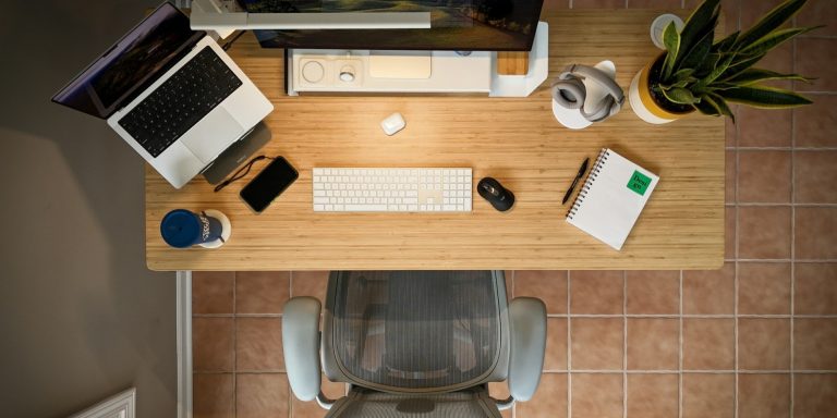 Top-down view of home office setup with a wooden desk, laptop, monitor, plant, and ergonomic mesh chair.
