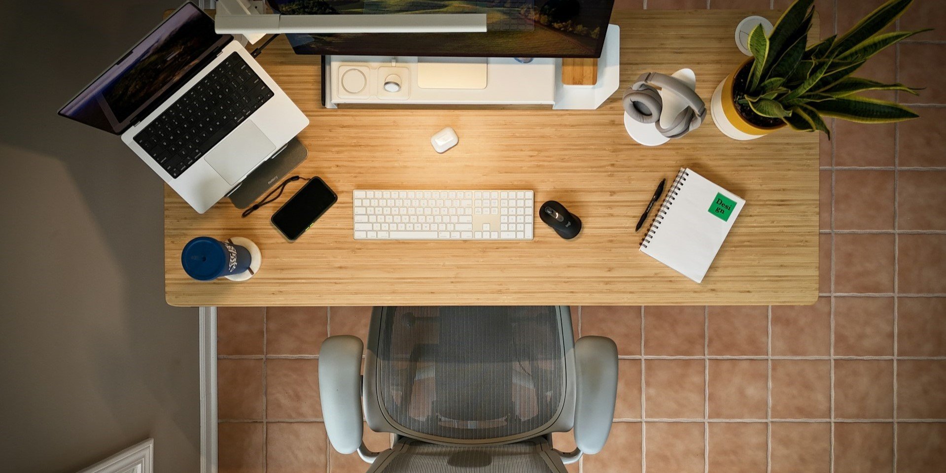 Top-down view of home office setup with a wooden desk, laptop, monitor, plant, and ergonomic mesh chair.